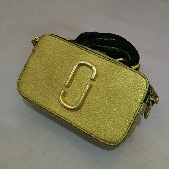 MARC JACOBS Snapshot Camera Bag METALLIC GOLD (Category: Luxury Bags & Wallets) located in Lucena. Submitted by brkrobert (Listing ID 1440582515, Image 2). Description: - Authentic Gold Marc Jacobs Snapshot camera bag - Features a detachable strap with black, white, and gold stripes - Strap is adorned with stylish metal stud details - All hardware is gold-tone - Shows some signs of wear on the hardware and strap, adding character.