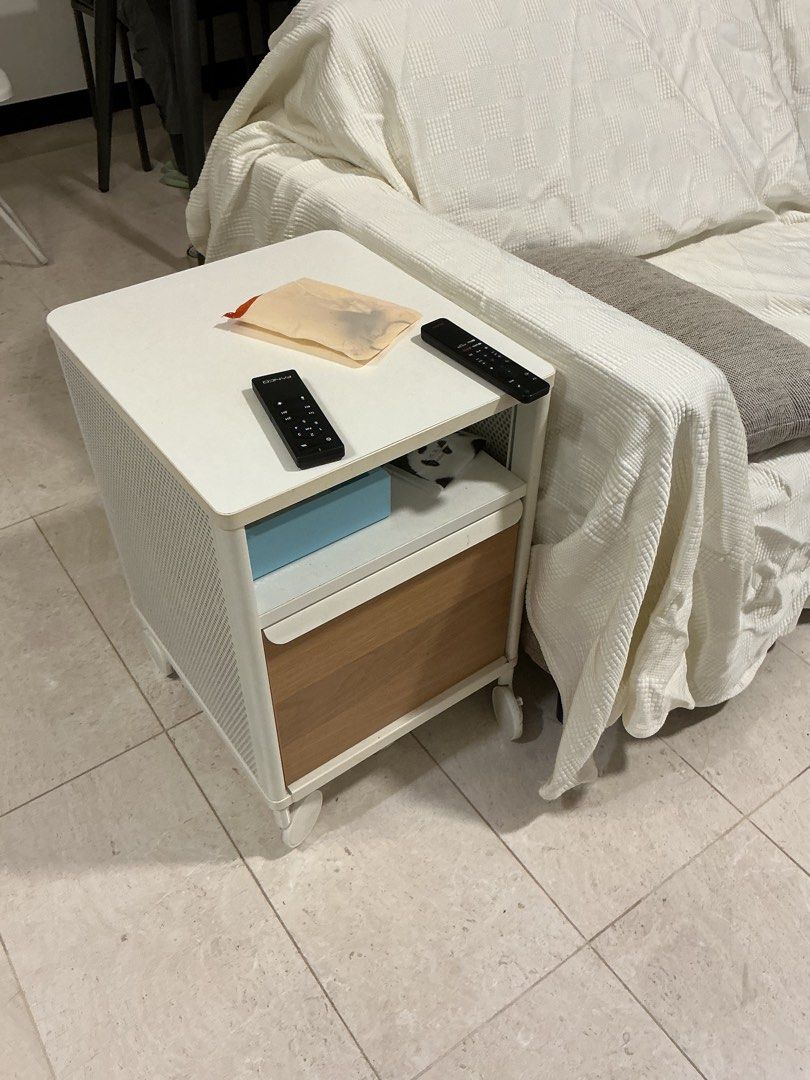 Side Table (Category: Tables & Sets) located in Singapore. Submitted by vantage.collective (Listing ID 1440774047, Image 1). Description: - White side table - Features a wooden drawer - Perforated design on the sides - Mounted on wheels for easy mobility - Ideal for use beside a sofa or bed - Offers generous surface and drawer space - Comes with two remotes and a small pouch on top.