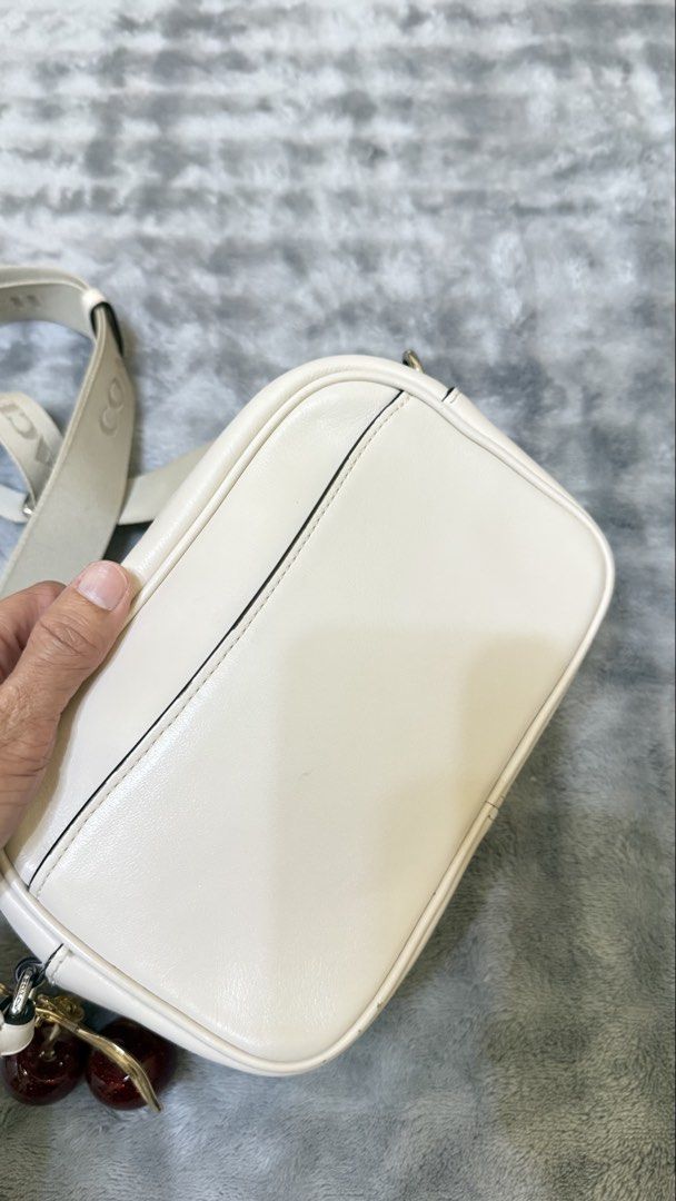 Coach Camera Bag with Cherry Charm (Category: Luxury Bags & Wallets) located in Bato. Submitted by babylaarni26255 (Listing ID 1436171678, Image 3). Description: - Cream-colored Coach camera bag - Detachable crossbody strap - Silver Coach logo - Cherry charm - Rectangular shape with zipper closure - Coach logo on strap - Includes tag.