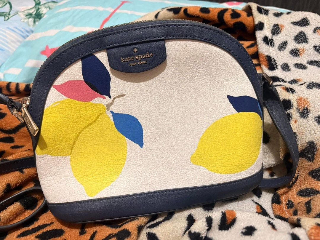 Kate Spade Lemon Print Crossbody Bag (Category: Cross-body Bags) located in Manila. Submitted by catherine46746 (Listing ID 1436450699, Image 1). Description: Kate Spade crossbody bag features a vibrant lemon print on a white background with navy blue trim. It has a zip closure and a navy blue strap. The bag is adorned with a Kate Spade New York logo. The design includes yellow lemons, pink and blue leaves, and a navy blue border. No flaws. .