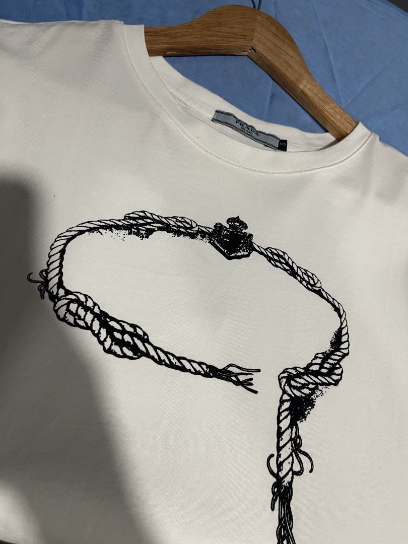 Prada Milano Rope Graphic T-shirt (Category: Tshirts & Polo Shirts ) located in Maasin. Submitted by niconii090 (Listing ID 1436297366, Image 2). Description: - Prada Milano T-shirt - Color: White - Graphic: Black rope design - ✅ 9.5/10 condition 📏20x25(inches) - Features: Crew neck, short sleeves - Condition: Excellent - Authentic Prada.