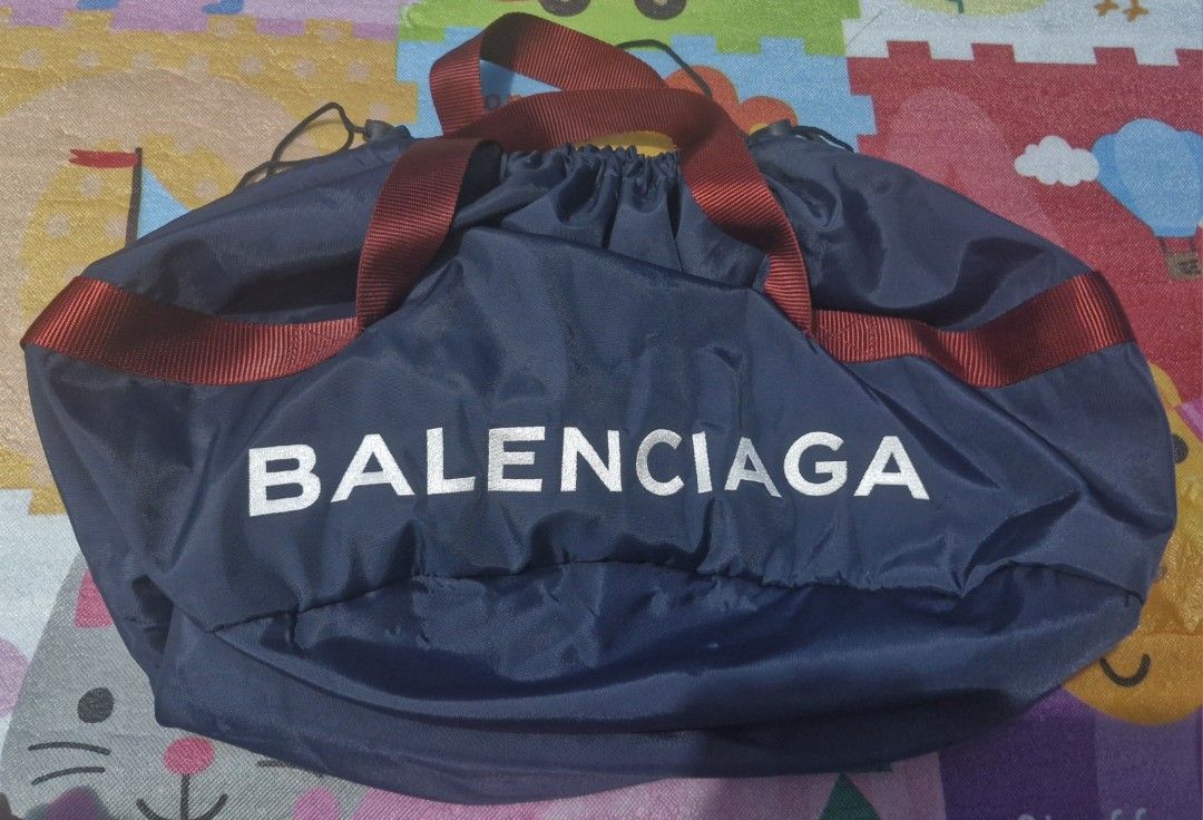 Balenciaga wheel duffle bag (Category: Luxury Bags & Wallets) located in Pili. Submitted by the_g_thrift (Listing ID 1436603192, Image 1). Description: - Balenciaga navy blue drawstring tote bag - Red strap and white Balenciaga logo - Drawstring closure - Circular base - Interior tag with brand name and "Paris" in gold - Serial number tag 19 diameter 14 length .