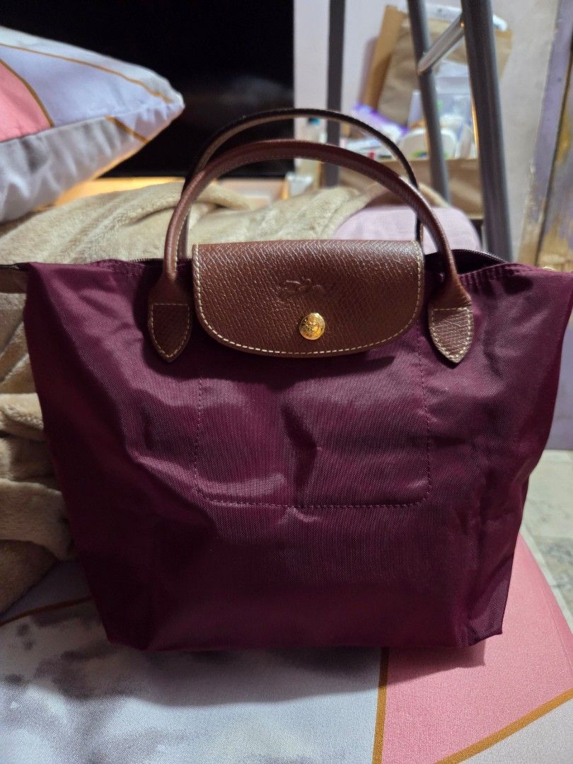 Longchamp Le Pliage Handbag - Burgundy (Category: Luxury Bags & Wallets) located in Makati City. Submitted by ellabit4379213 (Listing ID 1436921503, Image 3). Description: - Authentic Longchamp Le Pliage handbag - Color: Burgundy - Features brown leather handles and flap - Gold-tone button closure - Rectangular shape when folded - Made of a nylon-like material - Rounded, stitched handles - Simple, classic design.