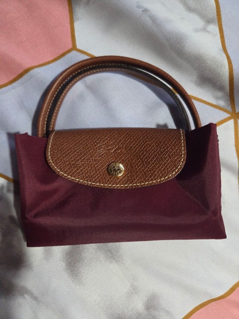 Longchamp Le Pliage Handbag - Burgundy (Category: Luxury Bags & Wallets) located in Makati City. Submitted by ellabit4379213 (Listing ID 1436921503, Image 2). Description: - Authentic Longchamp Le Pliage handbag - Color: Burgundy - Features brown leather handles and flap - Gold-tone button closure - Rectangular shape when folded - Made of a nylon-like material - Rounded, stitched handles - Simple, classic design.