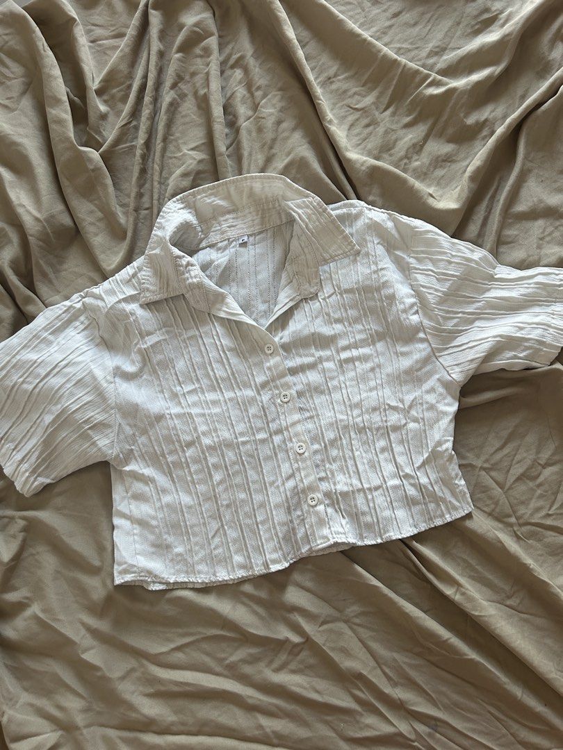 White Pleated Crop Top (Category: Blouses) located in Binan City. Submitted by missrandomthings (Listing ID 1436854609, Image 1). Description: White pleated crop top with short sleeves and a collared neckline. Features a button-down front closure. The fabric has a textured, pleated design. The top is a cropped length, suitable for pairing with high-waisted bottoms..