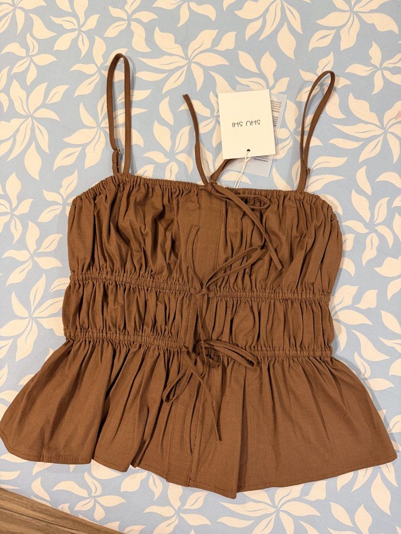 SHU SHI Brown Strap Top Linen Muse Top (Category: Sleeveless) located in Imus. Submitted by yummyfinds (Listing ID 1441302929, Image 1). Description: - Cute brown smocked top from SHU SHI - Features a tie front detail - Flattering peplum hem - Adjustable spaghetti straps - Made in Vietnam - Versatile: pairs well with white linen pants or jeans - Ideal for casual days or beach vacations.