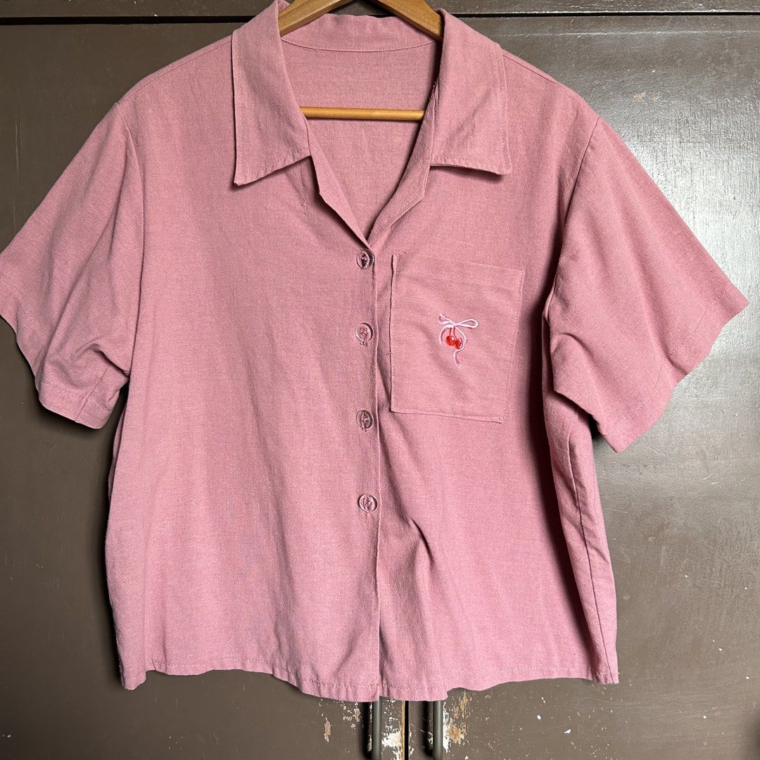 Pink Button-Up Shirt with Cherry Embroidery (Category: Shirts) located in Antipolo. Submitted by wonder.walls (Listing ID 1441430305, Image 1). Description: - Cute pink short-sleeved shirt - Features a pocket on the left chest - Adorned with sweet cherry and ribbon embroidery - Collared neckline with button closure - Relaxed fit for comfortable wear - Ideal for casual occasions.