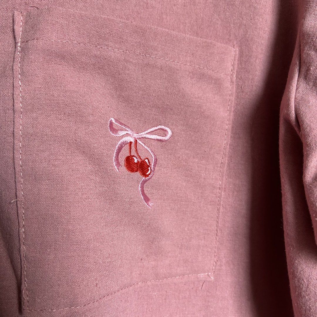 Pink Button-Up Shirt with Cherry Embroidery (Category: Shirts) located in Antipolo. Submitted by wonder.walls (Listing ID 1441430305, Image 2). Description: - Cute pink short-sleeved shirt - Features a pocket on the left chest - Adorned with sweet cherry and ribbon embroidery - Collared neckline with button closure - Relaxed fit for comfortable wear - Ideal for casual occasions.