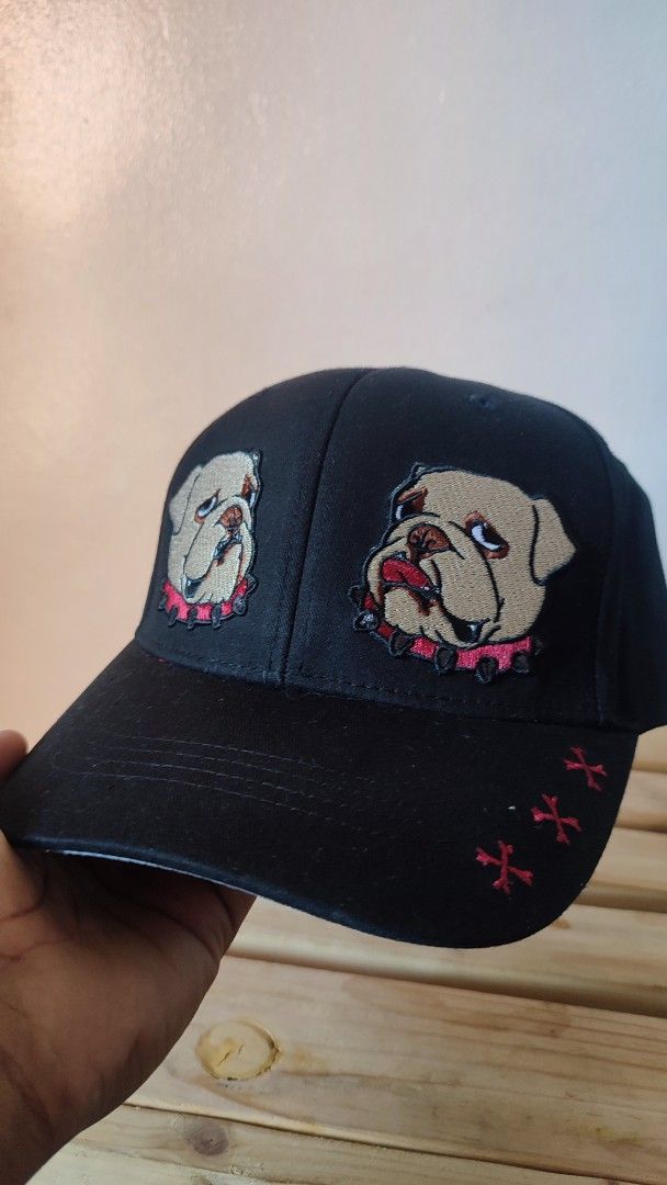 Black Baseball Cap with Bulldog Embroidery (Category: Caps & Hats) located in Manila. Submitted by janyvony_315 (Listing ID 1445753616, Image 2). Description: - Black baseball cap - Embroidered design: Two bulldog faces with pink spiked collars on the front - Embroidered red cross-like symbols on the side of the brim - White cross symbols on the back - Adjustable strap for a comfortable fit - Camouflage pattern on the underside of the brim.