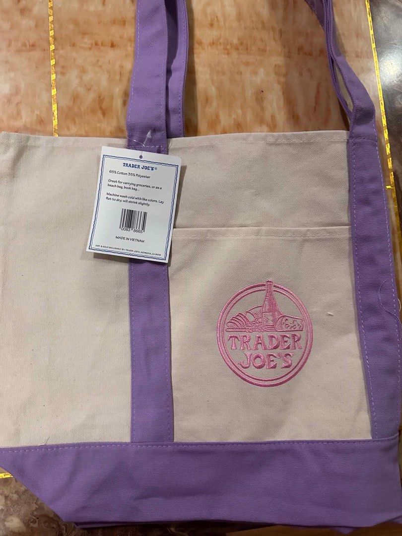 Trader Joe's Reusable Graphic Tote Bag (Category: Tote Bags) located in San Antonio. Submitted by pixiechromzzz (Listing ID 1446685003, Image 2). Description: - Trader Joe's Canvas Tote Bag from CALIFORNIA, USA - Color: Cream with purple accents - Features embroidered Trader Joe's logo on front - Includes a convenient front pocket - Material: 60% Cotton, 20% Polyester - Ideal for grocery shopping or everyday errands - Made in Vietnam.