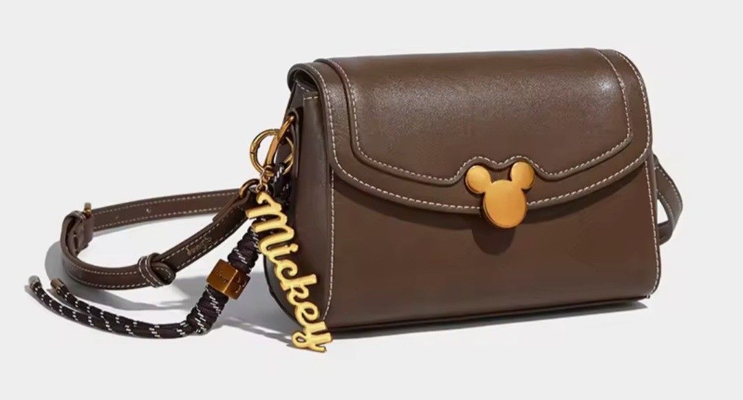 Brown Disney Mickey Mouse Crossbody Bag (Category: Cross-body Bags) located in Quezon City. Submitted by mimil_ (Listing ID 1441790493, Image 2). Description: - Cute brown crossbody bag - Features a Mickey Mouse clasp and "Mickey" charm - Comes with a stylish patterned strap - Perfect for everyday use or a fun Disney trip - Small and compact, ideal for carrying essentials - Detailed stitching on the flap -Bought at Laz*da Mall Disney Fashion Stor
