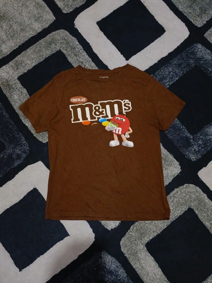 Brown M&M's Graphic T-Shirt (Category: Tshirts & Polo Shirts ) located in Andop. Submitted by kyutoryuu (Listing ID 1449076189, Image 1). Description: M&M's Tee •Dimes: 17.5x24 •Condition: As New •CR: 9.8/10 •Choco Brown •Issue: need wash.