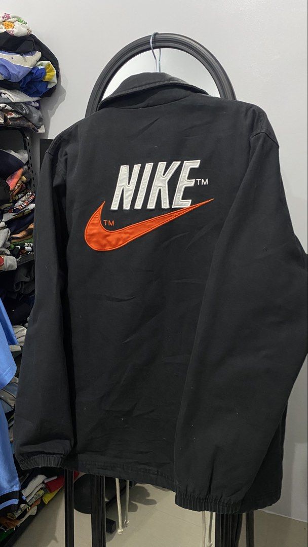 Nike Black Coach Jacket with Swoosh Logo (Category: Coats, Jackets and Outerwear) located in Porac. Submitted by moethrifter (Listing ID 1449167059, Image 1). Description: - Classic black Nike coach jacket - Iconic orange swoosh and Nike logo embroidered on back - Quilted lining for added warmth - Snap button closure - Two side pockets - Ideal for everyday wear.