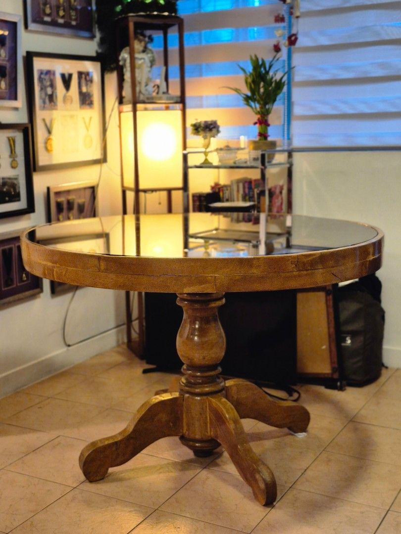 Round Solid Wood Table with Glass Top (Category: Tables & Sets) located in Makati City. Submitted by femosseverything (Listing ID 1451182964, Image 2). Description: - Round dining table with a solid wood base and a clear glass top. - Features a classic, sturdy pedestal leg design. - Warm, natural wood finish. - Condition: Used, with a minor chip on the wood edge (refer to the last image). - Ideal for dining areas or as a decorative accent. - Dime