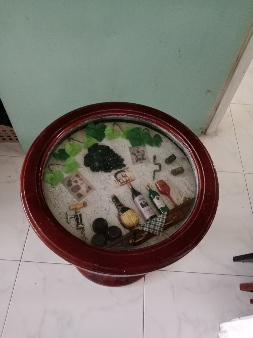 Round table (Category: Tables & Sets) located in Singapore. Submitted by janetsklim (Listing ID 1451154856, Image 3). Description: - Unique round table with a dark wood finish. - Features a decorative glass top with a detailed wine-themed design. - Approximate dimensions: 18.5" diameter, 18.5" height. - Adds a touch of rustic elegance to any room. - In good, pre-loved condition..
