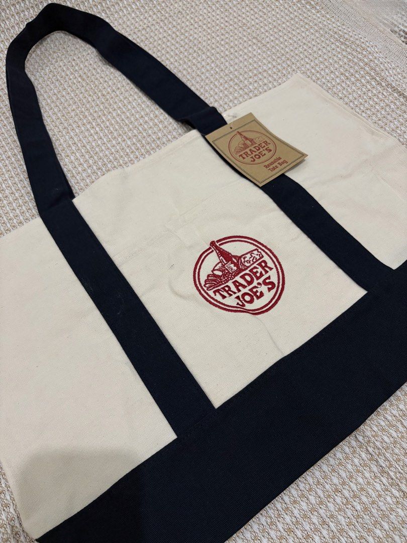 Trader Joe's Large Canvas Tote Bag (Category: Tote Bags) located in Manila. Submitted by love.loop (Listing ID 1452729152, Image 2). Description: - Classic Trader Joe's reusable tote bag - Cream color with black handles and base - Features iconic red embroidered Trader Joe's logo - Durable canvas material - Perfect for groceries or everyday use.