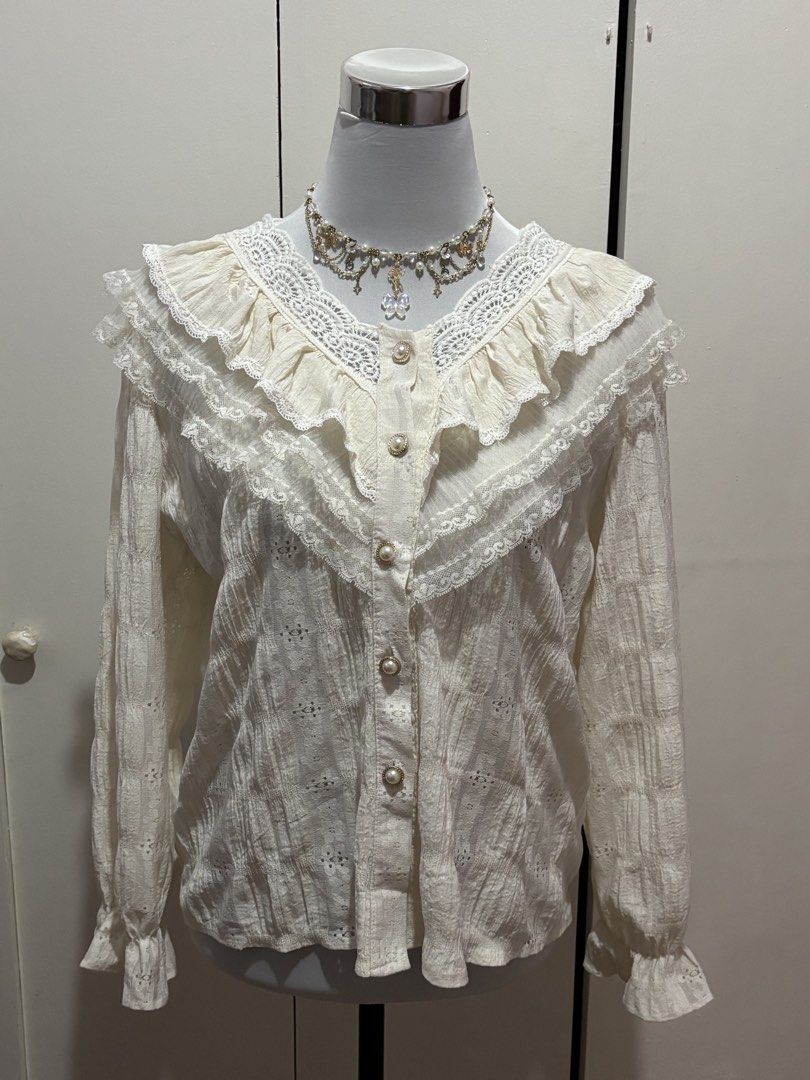 (m-l) White Lace Ruffle Long Sleeve Blouse (Category: Blouses) located in Paranaque City. Submitted by prelovedtinez (Listing ID 1448042393, Image 1). Description: - Cream white blouse with intricate lace details - Features ruffles along the neckline and sleeves - Button-up front with pearl-like buttons - Textured fabric with eyelet details - Long sleeves with ruffled cuffs - Delicate and vintage-inspired design.