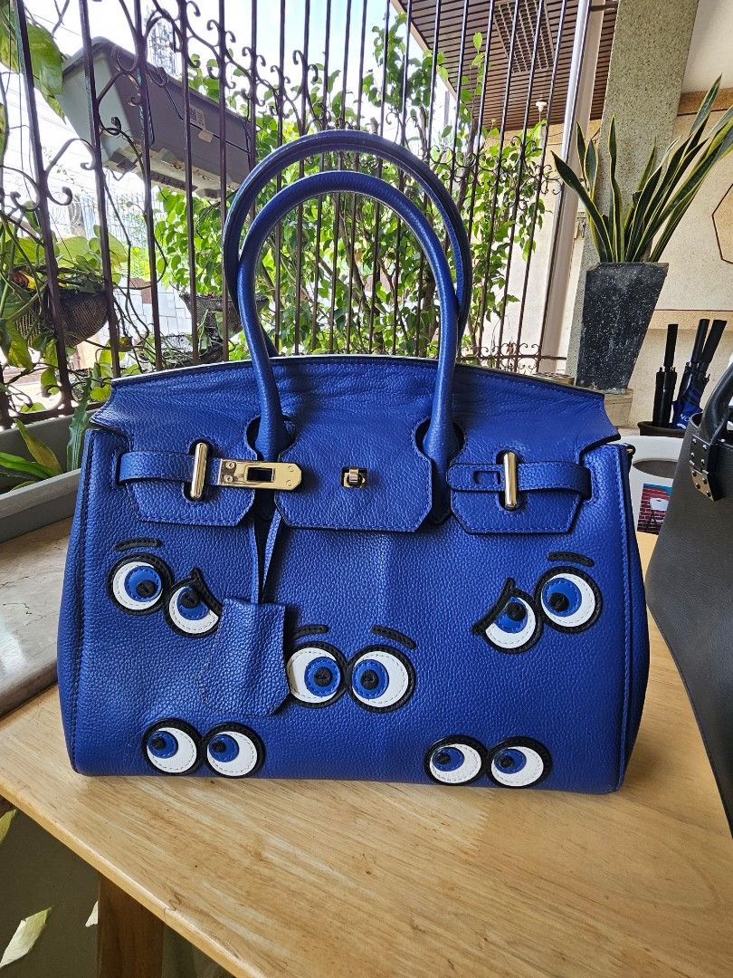 Blue Handbag with Googly Eyes Design (Category: Luxury Bags & Wallets) located in Cabanatuan. Submitted by eclaudio (Listing ID 1448610710, Image 1). Description: - Fun blue handbag with unique googly eyes design. - Classic structured shape. - Features top handles and a secure clasp closure. - Perfect statement piece for quirky accessory lovers. - Spacious enough to hold your daily essentials. - Ready for its new home!.