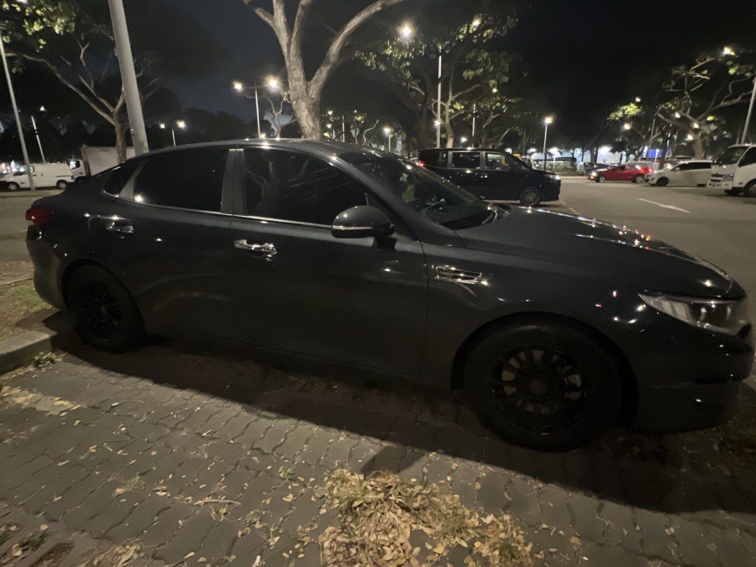 Kia Optima K5 2.0 (A) Auto (Category: Used Cars) located in Singapore. Submitted by v_random (Listing ID 1448660591, Image 2). Description: Dark grey Kia sedan. Front view of the car. Grille and headlights are visible. Parked outdoors at night. Some reflections on the windshield. Well maintained and accident-free. Original paintwork. Meet-up for viewing available Left 3 months +.