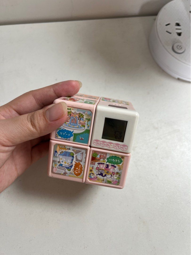 🎲WanTame Hapi Hapi Cube Town (Category: Interactive Toys) located in Tarlac. Submitted by just_decluttering (Listing ID 1455915192, Image 2). Description: 🎲ORIGINAL PRICE: ₱4,549.53 -w/o box -Release by Takara Tomi - Adorable Tamagotchi cube toy - Features a black and white screen - Offers various game screens and characters - Comes in a pink casing with colorful illustrations - Ideal for collectors and fans of retro digital pets.