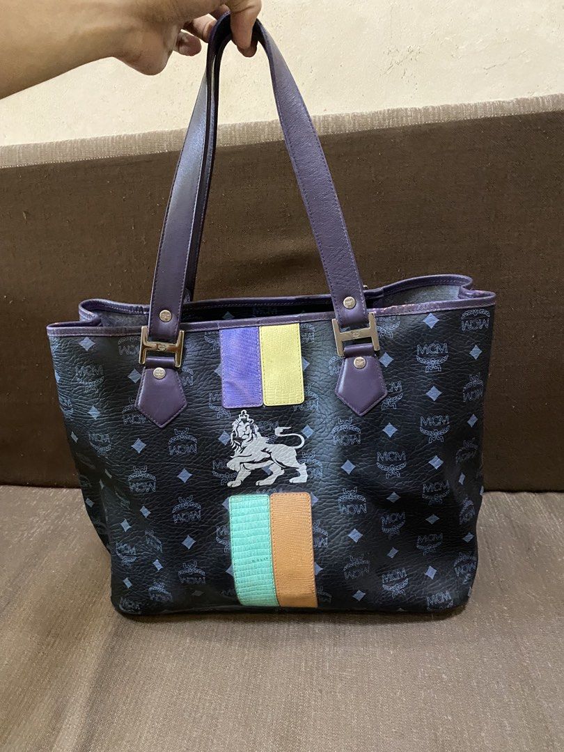 MCM Tote Bag with Lion Emblem (Category: Luxury Bags & Wallets) located in San Antonio. Submitted by pixiechromzzz (Listing ID 1456128110, Image 1). Description: - Authentic MCM tote bag in black with signature pattern. - Features a striking central lion emblem. - Adorned with colorful stripes above and below the emblem. - Elegant purple leather trim and handles. - Polished gold hardware details. - Spacious interior, ideal for everyday use..