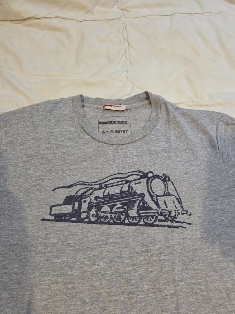 Prada Train Engine Graphic Tee (Category: Tshirts & Polo Shirts ) located in Taguig. Submitted by mightydept (Listing ID 1456136614, Image 3). Description: - Grey Prada T-shirt - Features a cool engine graphic print on the front - Prada logo and article number clearly visible on tag - Wash tag faded; Fits Medium-Large Mens.