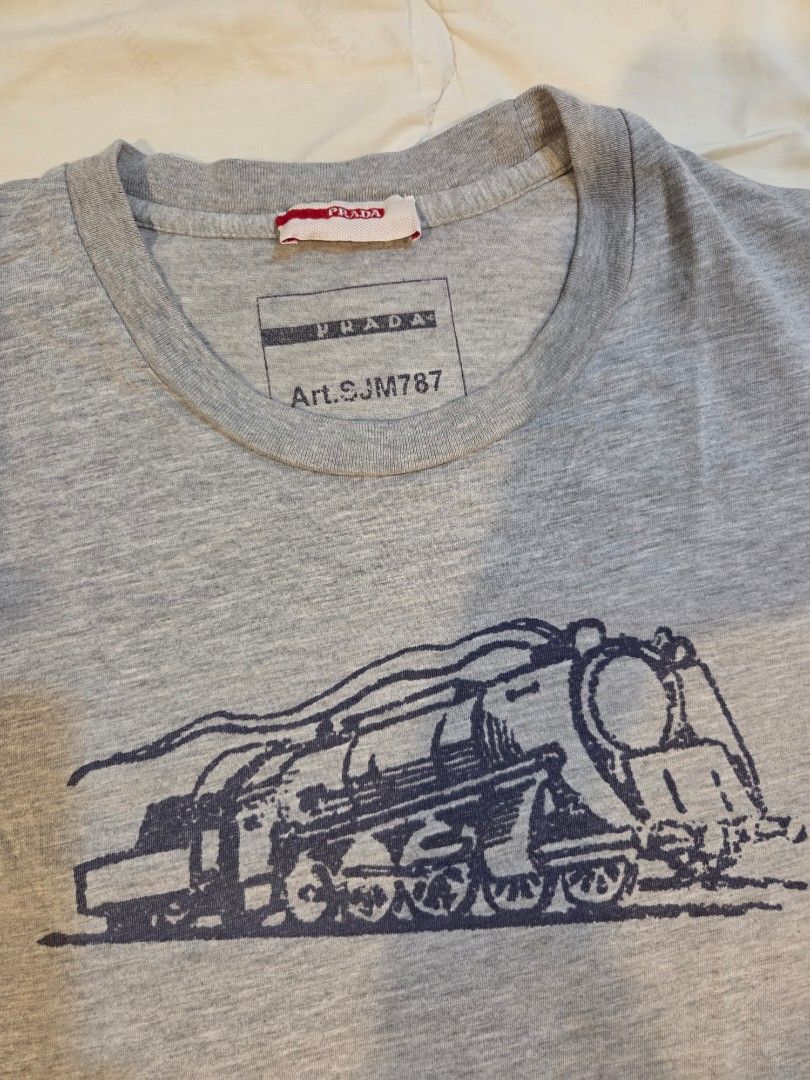 Prada Train Engine Graphic Tee (Category: Tshirts & Polo Shirts ) located in Taguig. Submitted by mightydept (Listing ID 1456136614, Image 2). Description: - Grey Prada T-shirt - Features a cool engine graphic print on the front - Prada logo and article number clearly visible on tag - Wash tag faded; Fits Medium-Large Mens.