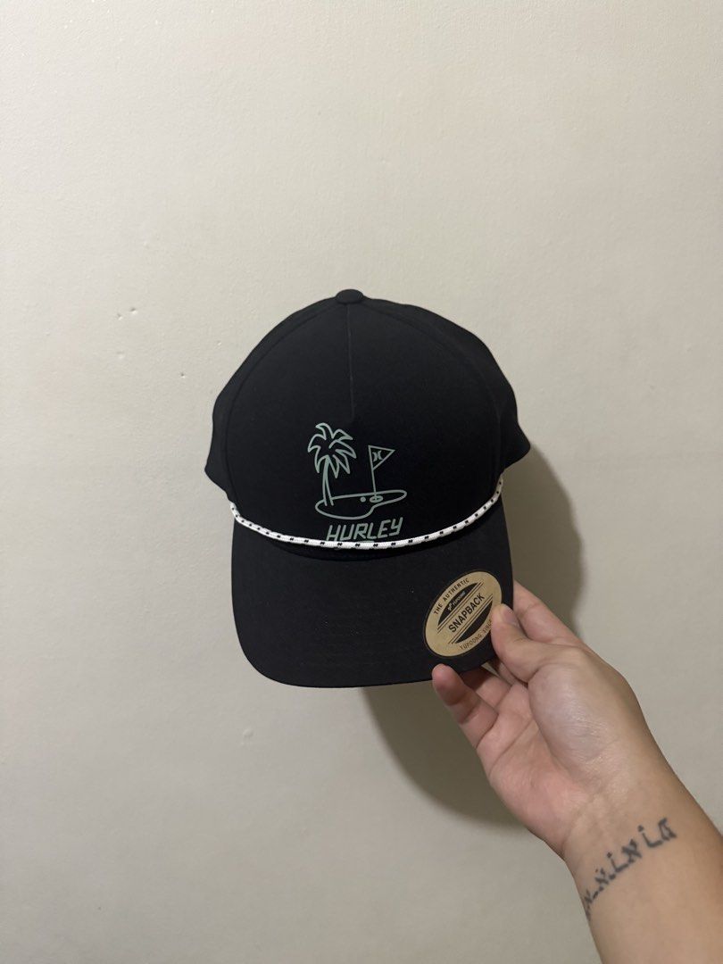 Hurley Black Snapback Cap with Rope Detail (Category: Caps & Hats) located in Manila. Submitted by mrtndmnc (Listing ID 1453401488, Image 1). Description: - Black Hurley snapback cap - Rope detail across the brim - Embroidered palm tree and golf flag graphic on the front - Perforated sides for enhanced breathability - Adjustable snapback closure for a custom fit - Ideal for everyday casual wear.