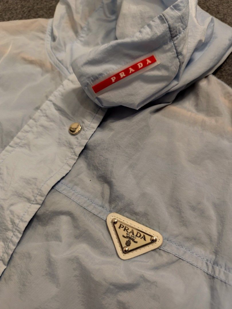 Prada Light Blue Hooded Windbreaker Jacket (Category: Luxury Apparel) located in Cordova. Submitted by bsb.streetluxvintage.clothing (Listing ID 1457259530, Image 2). Description: - Light blue Prada windbreaker jacket - Hooded design with drawstring hem - Two front pockets - Zip-up closure - Iconic Prada triangle logo on chest - Ideal for casual wear or light outdoor activities.