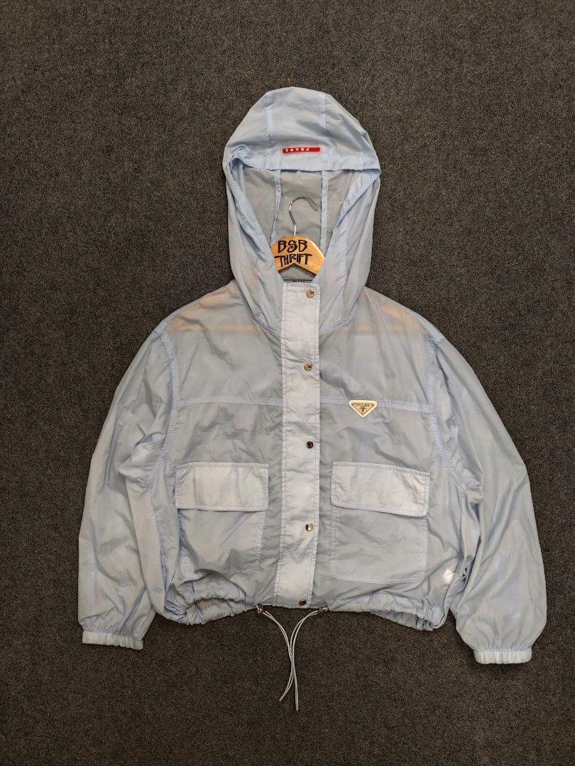 Prada Light Blue Hooded Windbreaker Jacket (Category: Luxury Apparel) located in Cordova. Submitted by bsb.streetluxvintage.clothing (Listing ID 1457259530, Image 1). Description: - Light blue Prada windbreaker jacket - Hooded design with drawstring hem - Two front pockets - Zip-up closure - Iconic Prada triangle logo on chest - Ideal for casual wear or light outdoor activities.