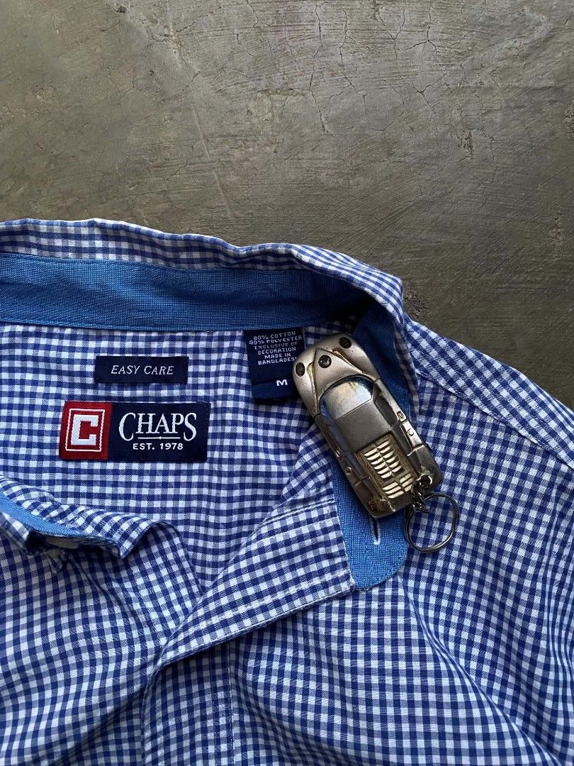 Chaps Easy Care Gingham Button Down Shirt (Category: Tshirts & Polo Shirts ) located in Cabanatuan. Submitted by vivian_nicole_03 (Listing ID 1453535791, Image 3). Description: - Classic blue and white gingham pattern - Brand: Chaps - Style: Button-down shirt - Neckline: Collared - Closure: Button - Features: Chest pocket with embroidered Chaps logo - Fabric: Easy care, suitable for everyday wear - Size: Medium (M).