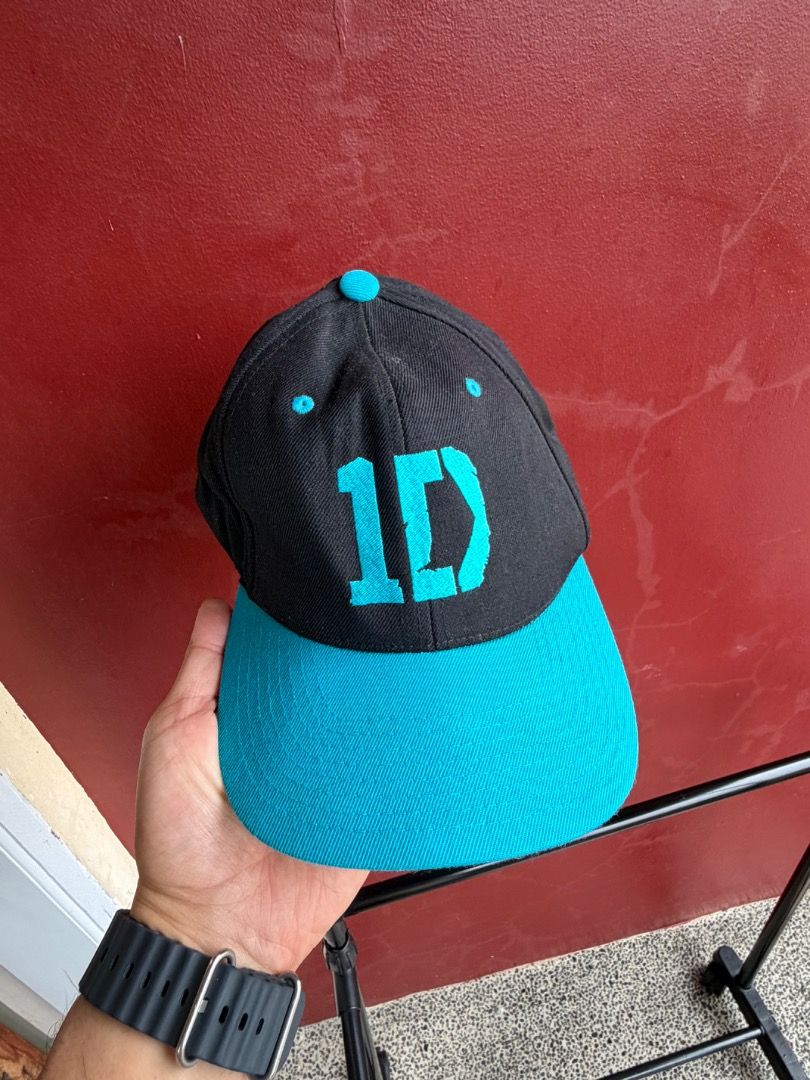 Official One Direction 1D Baseball Cap (Category: Caps & Hats) located in Manila. Submitted by transbox (Listing ID 1458545935, Image 1). Description: Colorway & Style: Black crown paired with a vibrant teal/turquoise flat brim, matching top button, embroidered eyelets, and snapback strap. Graphics: Features the bold 1D logo embroidered in teal on the front panel. Brand & Tag: Built on "The Classics Yupoong" base cap (80% acrylic, 20% wool; Made 