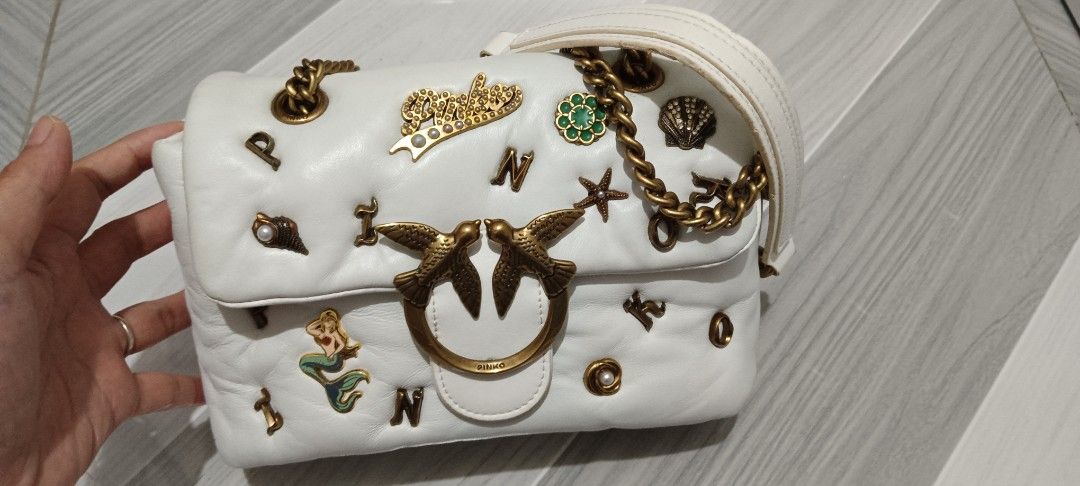 Pinko White Leather Bag with Charms (Category: Luxury Bags & Wallets) located in Abucay. Submitted by jazelyfindzshop (Listing ID 1458577203, Image 2). Description: - White leather crossbody bag by Pinko - Adorned with charming mermaid, crown, and pearl embellishments - Elegant gold chain strap - Signature Pinko swallow clasp closure - A unique and eye-catching accessory to elevate any look.