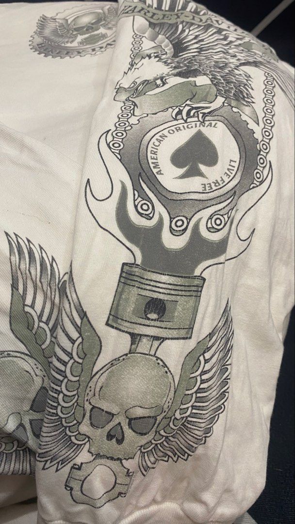 Harley-Davidson T-Shirt Manassas VA Eagle Skull Piston (Category: Tshirts & Polo Shirts ) located in Porac. Submitted by moethrifter (Listing ID 1458792315, Image 3). Description: - White Harley-Davidson T-shirt - Size: XL - Front Graphic: American flag, motorcycle, cannon, 'Whitts Harley-Davidson Manassas, VA' - Back Graphic: Large eagle with skull, piston, and flames - Design: Detailed and colorful.