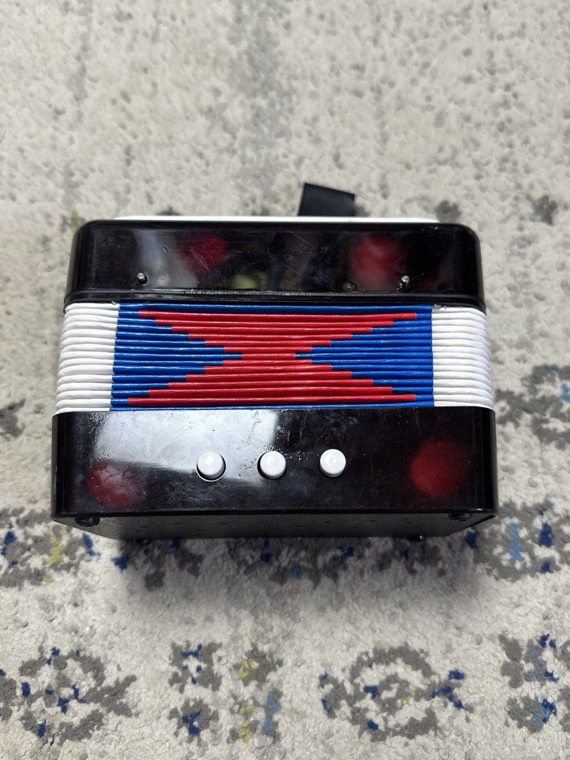 Mini Accordion Toy Musical Instrument (Category: Kids Toys) located in Daet. Submitted by thrifthings2018 (Listing ID 1458773365, Image 2). Description: - Black toy accordion, perfect for kids! - Vibrant red, white, and blue bellows for a fun look. - White buttons on top and bottom for easy play. - Includes a small strap on top. - Measures approximately 7 inches tall. - Great for sparking musical creativity!.