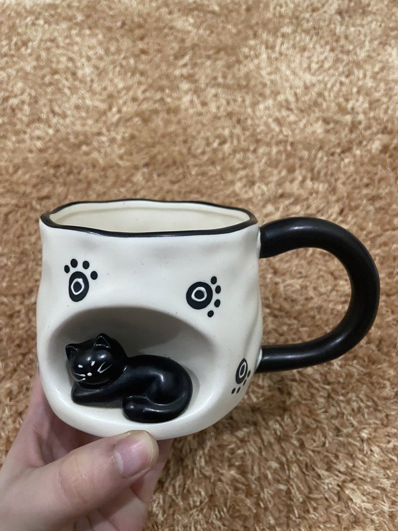 Cute Black Cat Ceramic Mug with Paw Prints (Category: Coffee & Tea Tableware) located in Guiguinto. Submitted by beatan13 (Listing ID 1453766216, Image 1). Description: - Adorable white ceramic mug - Features a unique cutout of a sleeping black cat - Decorated with black paw print details - Includes a black handle - Ideal gift for cat lovers - Perfect for enjoying your morning coffee or tea.