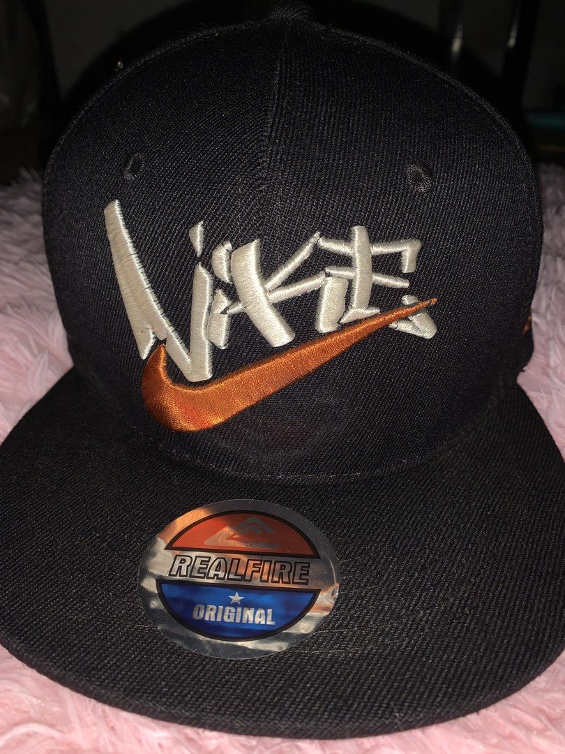 Nike Snapback Cap - Black with Orange Swoosh (Category: Caps & Hats) located in Binan City. Submitted by kariray_shop (Listing ID 1454065965, Image 1). Description: - Classic black Nike snapback cap - Iconic orange Nike swoosh logo - 'NIKE' embroidered in orange on the side - Unique 'REAL FIRE' branding inside - Adjustable snap closure for a custom fit.