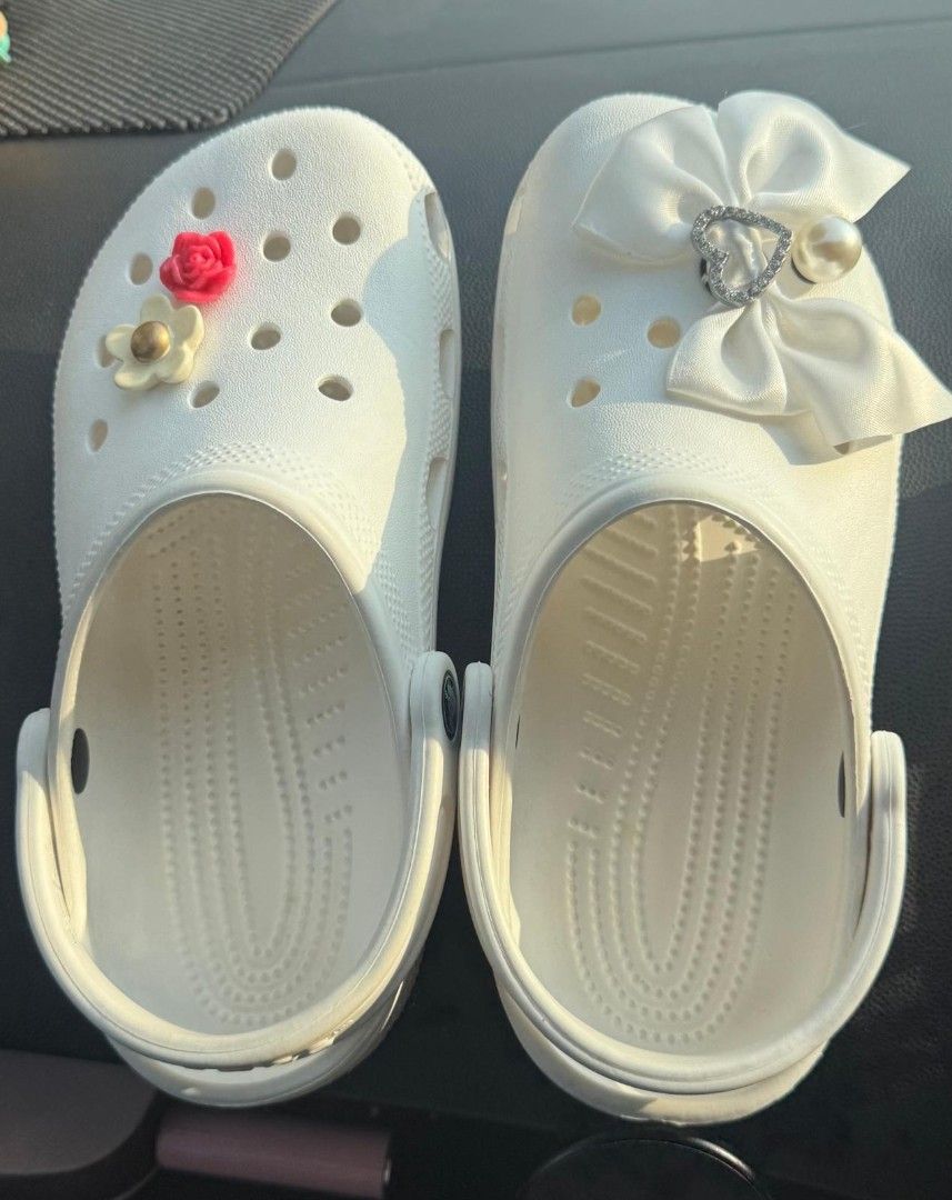Crocs (Authentic) (Category: Sandals) located in Singapore. Submitted by egwd. (Listing ID 1454563442, Image 1). Description: - SIZE J5 - White Crocs - Decorated with unique Jibbitz charms - One shoe features a red flower and gold accent - Other shoe adorned with a white bow, heart charm, and pearl - Classic Crocs comfort and style - Versatile white color.