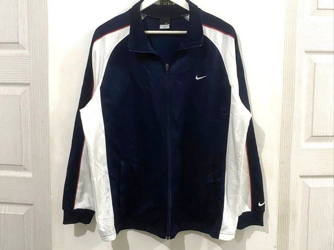 Nike Track Jacket (Category: Coats, Jackets and Outerwear) located in Bulacan. Submitted by wowee (Listing ID 1454576830, Image 1). Description: Navy blue Nike track jacket with white and red accents. Features a full zip closure and AE the Nike logo on the chest. The jacket has a ribbed collar and cuffs. The size tag indicates a Men's L. Made of polyester. Includes Japanese text on the tag..