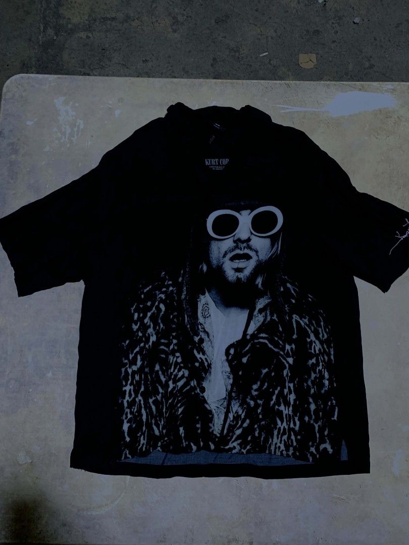 Kurt Cobain Graphic T-Shirt (Category: Tshirts & Polo Shirts ) located in Las Pinas. Submitted by akositroyvhaha (Listing ID 1461094903, Image 1). Description: - Black t-shirt with large Kurt Cobain graphic print - Features iconic sunglasses and leopard print jacket - 'KURT COP' brand visible on collar - Ideal for Nirvana fans.