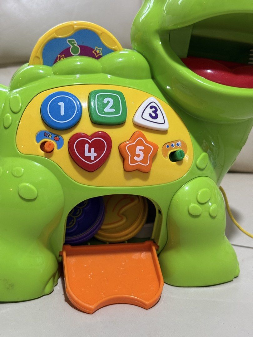 Educational toys VTech Chomp & Count Dino (interactive learning toys) (Category: Infant Playtime) located in Angeles City. Submitted by holyfabclothing (Listing ID 1460938661, Image 2). Description: VTech Chomp & Count Dino an interactive learning toy for toddlers featuring a green electronic dinosaur with colorful numbered buttons and a set of plastic food pieces. Toy Features Interactive Feeding: The dinosaur recognizes different colored food p