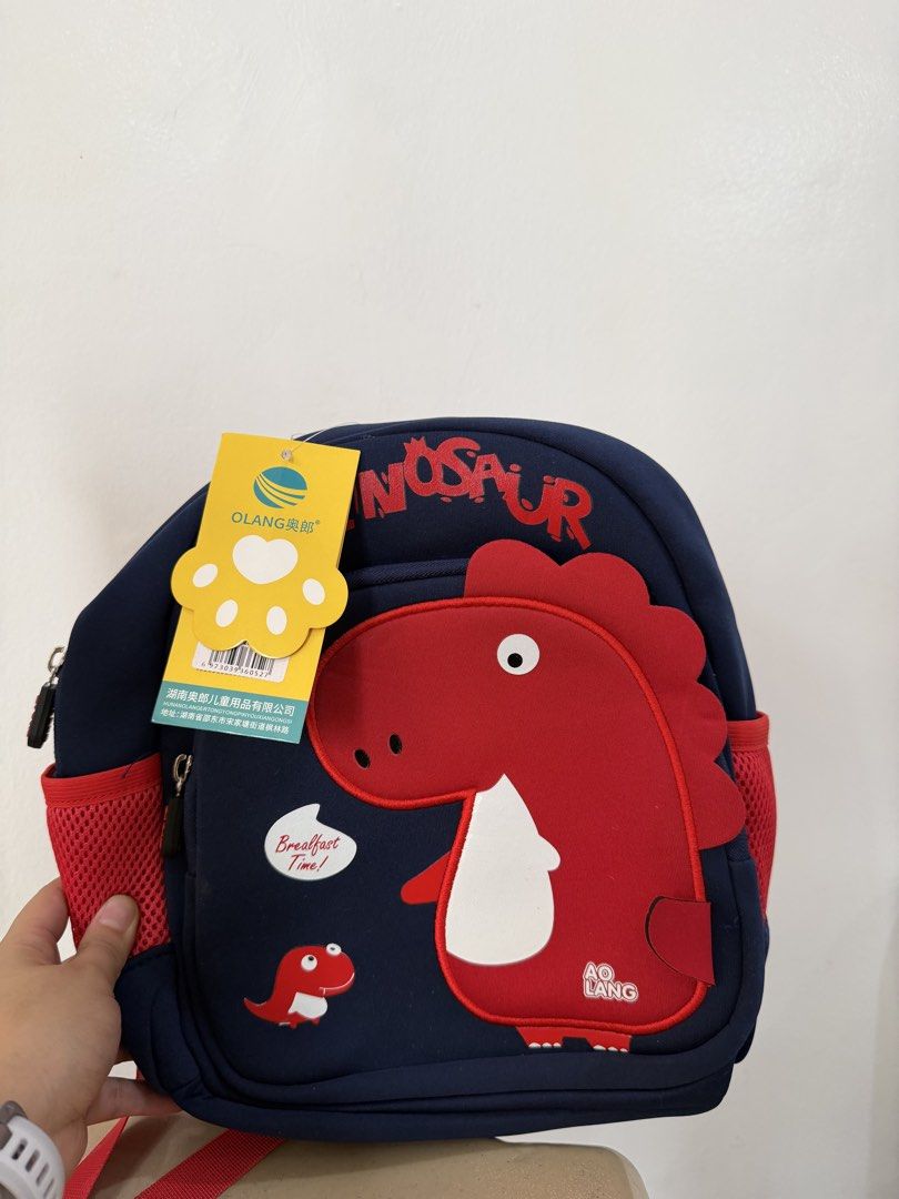 Olang Dinosaur Backpack for Kids (Category: Babies & Kids Fashion) located in Manila. Submitted by escarchame98247 (Listing ID 1461161979, Image 1). Description: - Cute navy blue Olang backpack - Features a big red dinosaur design ('NOSAUR') - Includes 'Breakfast Time!' graphic with a smaller dinosaur - Convenient mesh side pockets for water bottles - Ideal for school, daycare, or everyday outings.