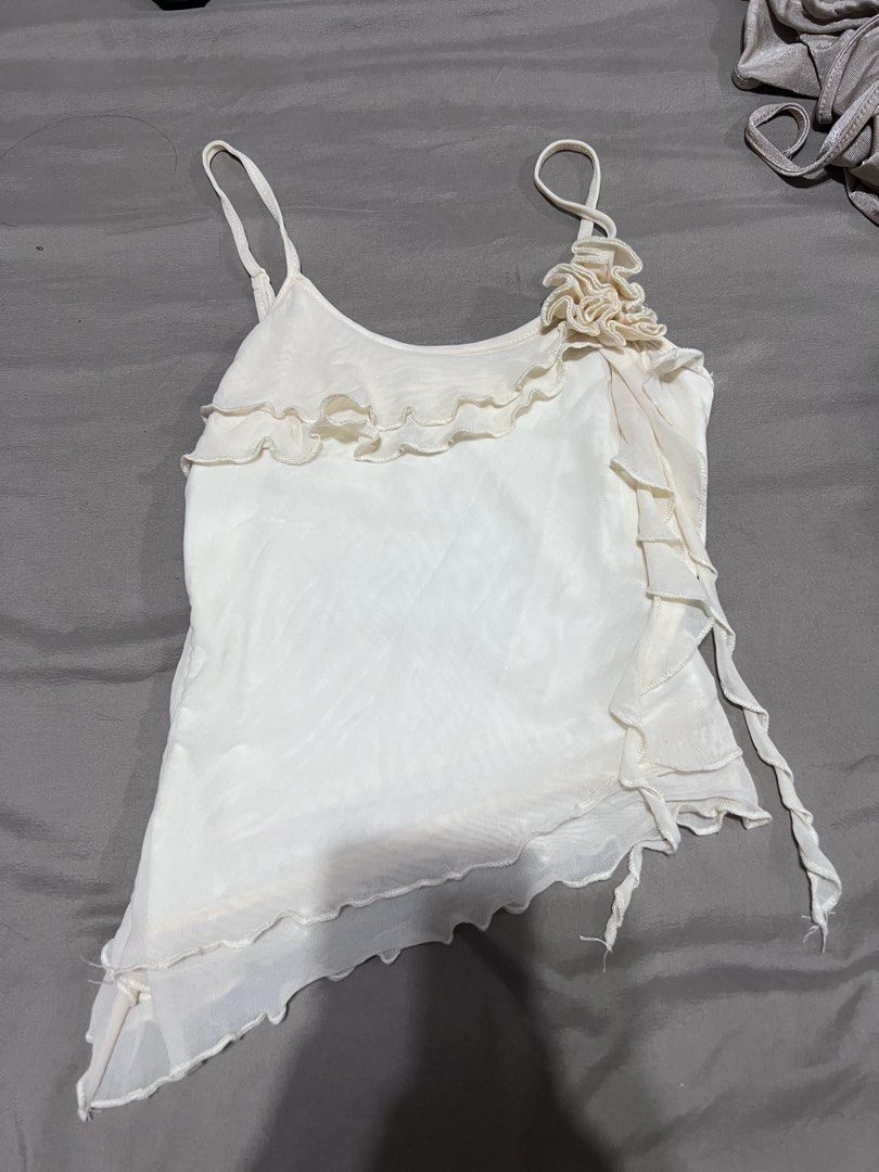 White Ruffled Cami Top with Flower Detail (Category: Sleeveless) located in Quezon City. Submitted by jinnysells (Listing ID 1461276458, Image 1). Description: - Adorable white cami top - Features charming ruffles on neckline and hem - Pretty flower detail on the strap - Unique asymmetrical hemline with sheer fabric - Versatile for layering or wearing on its own - Creates a sweet and feminine aesthetic.