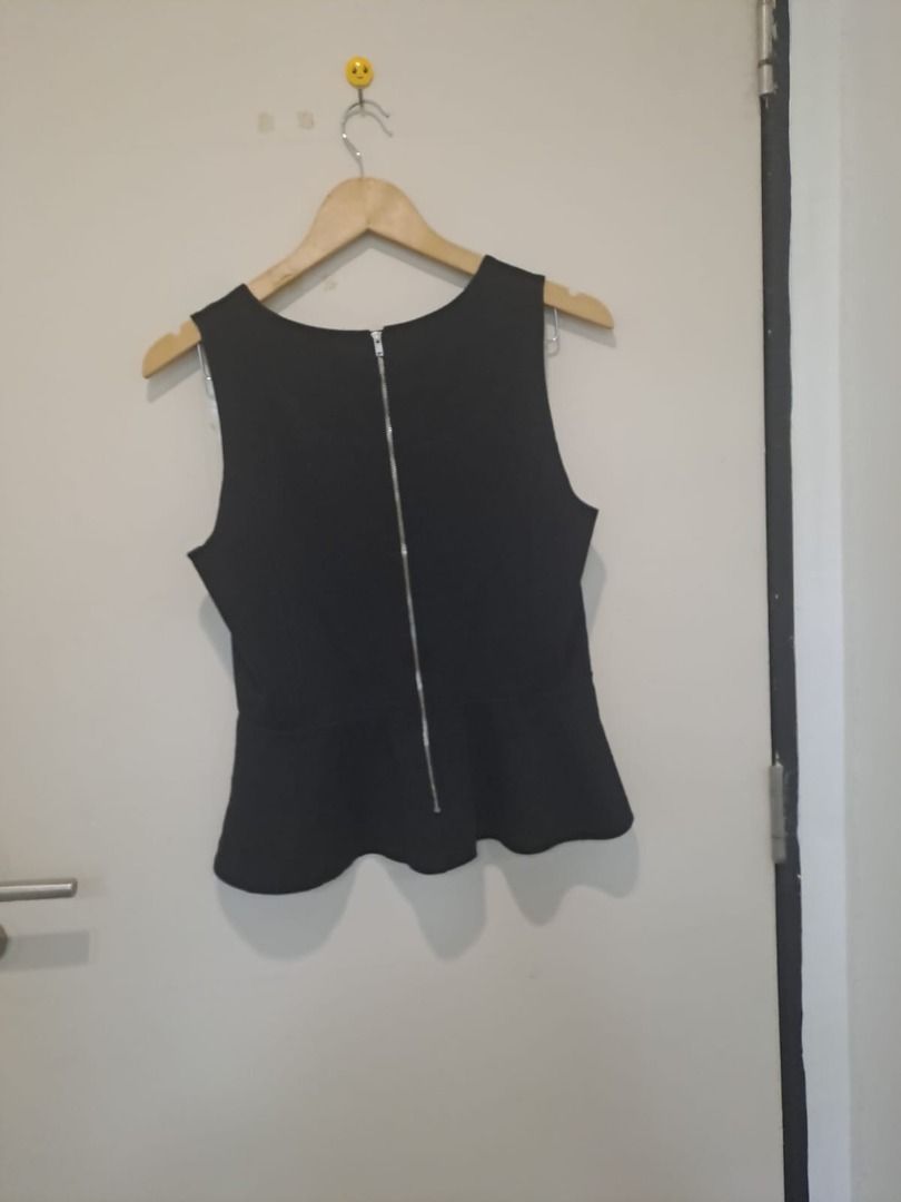 peplum black sleeveless (Category: Sleeveless) located in Mandaluyong City. Submitted by epicowl-c93c (Listing ID 1461350854, Image 3). Description: The image shows a black sleeveless peplum top hanging on a wooden coat hanger against a white door. The top features a crew-style round neckline, structured darting along the bodice for a fitted silhouette, a seam at the waist, and a characteristic flared peplum hem.This timeless, versatile piece bri