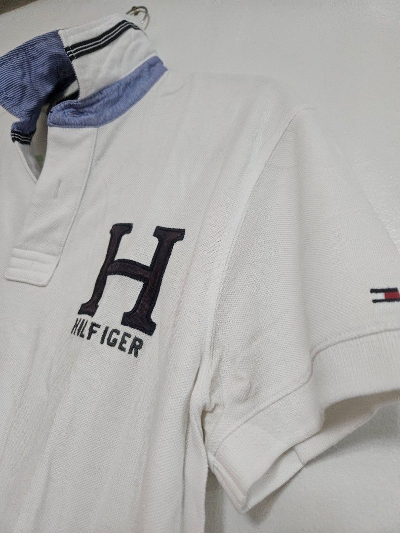 Tommy Hilfiger White Polo Shirt (Category: Tshirts & Polo Shirts ) located in Batangas. Submitted by golem08 (Listing ID 1461405556, Image 2). Description: - Classic white Tommy Hilfiger polo shirt - Large 'H' logo with 'Hilfiger' text on the chest - 'East Div 815 New York' graphic on the front - Features a classic collar and button placket - Short sleeves with ribbed cuffs - Ideal for casual everyday wear.