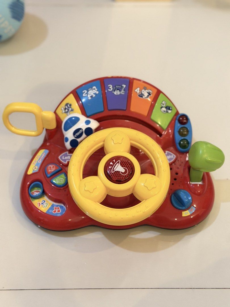 VTech Go! Go! Smart Wheels Steering Wheel Toy (Category: Kids Toys) located in Rizal. Submitted by karlaquijanomd (Listing ID 1461386539, Image 1). Description: - Interactive VTech steering wheel for pretend play. - Features numerous buttons for sounds and music. - Includes a horn, gear shifter, and traffic lights. - The car on top is branded 'VTech'. - Helps children learn numbers 1-4. - Requires batteries to operate..