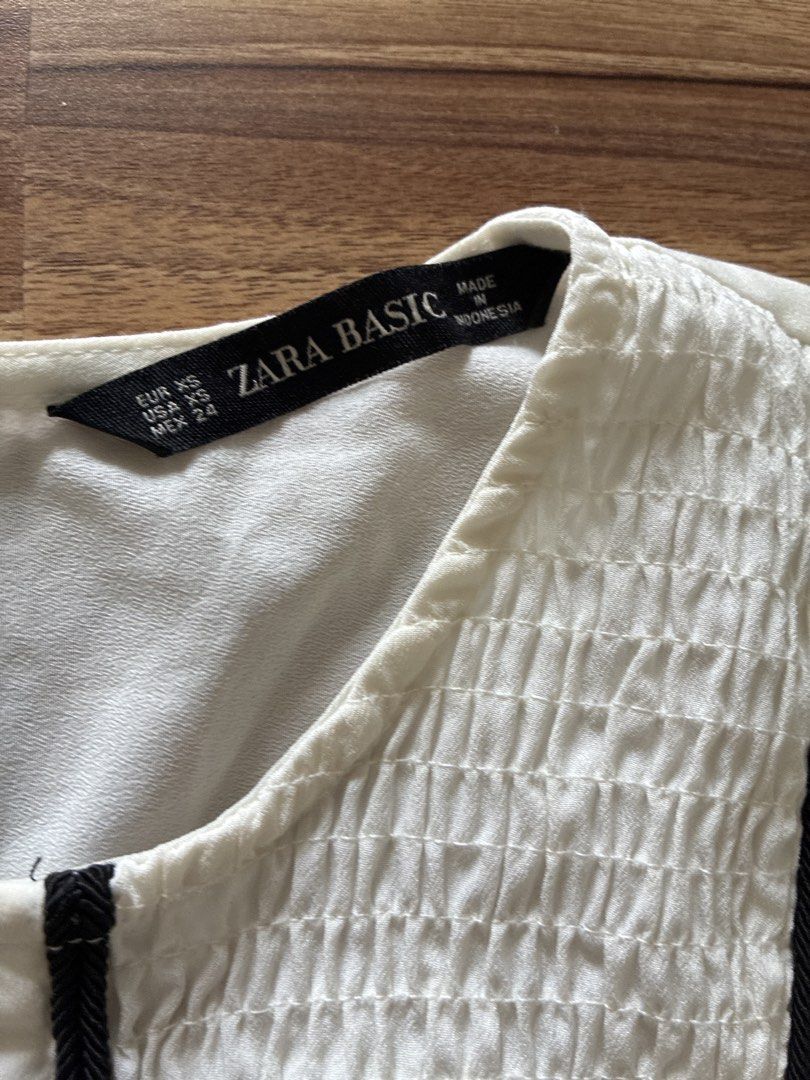 Zara Basic White Pleated Smocked Button Down Sleeveless Top XS (Category: Sleeveless) located in Pasig City. Submitted by slr1949 (Listing ID 1461409989, Image 2). Description: - White sleeveless top from Zara Basic - Smocked bodice with black button accents - Pleated design on the lower part - Black trim along neckline and down the front - Versatile for casual or dressed-up looks.