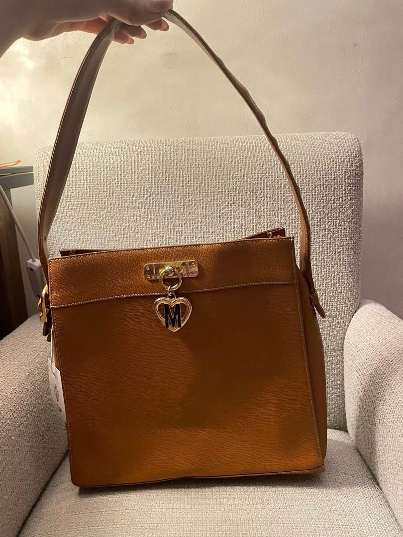 Moschino Leather Handbag with Heart Charm (Category: Luxury Bags & Wallets) located in Quezon City. Submitted by hadescloset (Listing ID 1461690877, Image 1). Description: - Authentic Moschino brown leather handbag - Features a gold-tone heart charm with 'M' initial - Top handle and detachable shoulder strap - Textured exterior finish - Secure zipper closure at the top - Comes with original dust bag - Ideal for everyday wear and adding a touch of