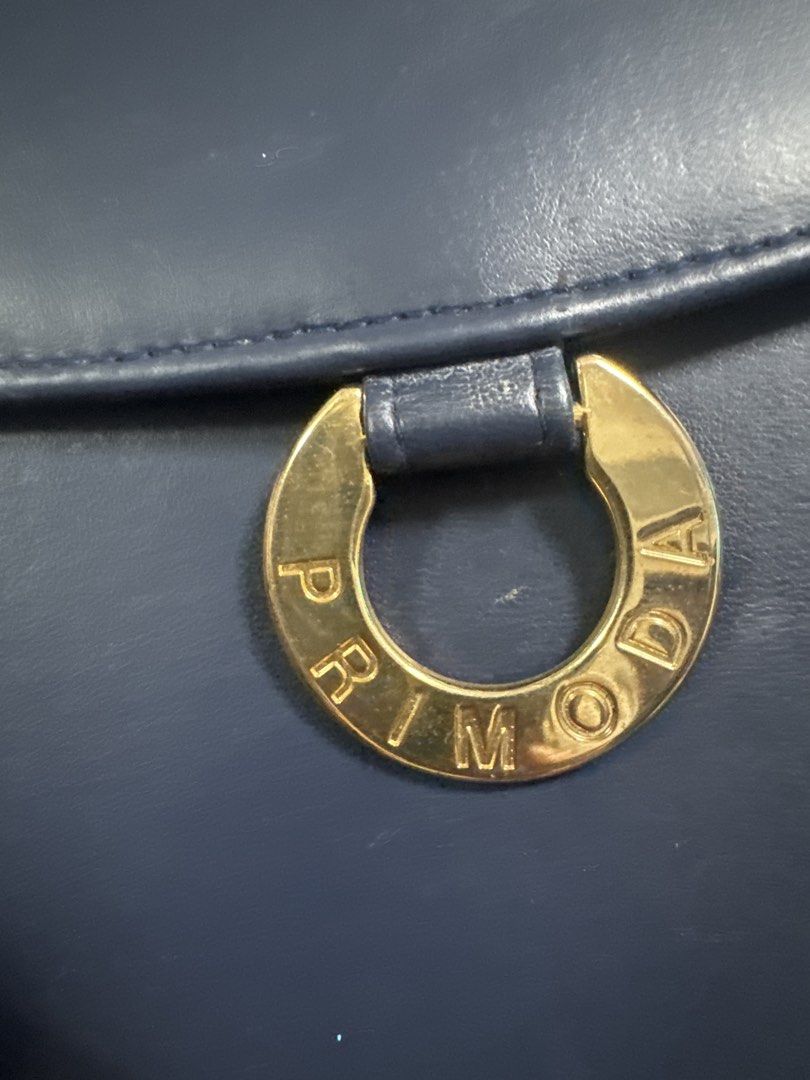 2027 PRIMOD A Navy Blue Leather Handbag (Category: Luxury Bags & Wallets) located in Silang. Submitted by sari2bilina (Listing ID 1462148617, Image 3). Description: - Navy blue leather handbag - Gold ring detail with 'PRIMOD A' embossed on front - Top handle - Clean interior - Approx. 8" tall x 6" wide - Versatile for everyday or special occasions.