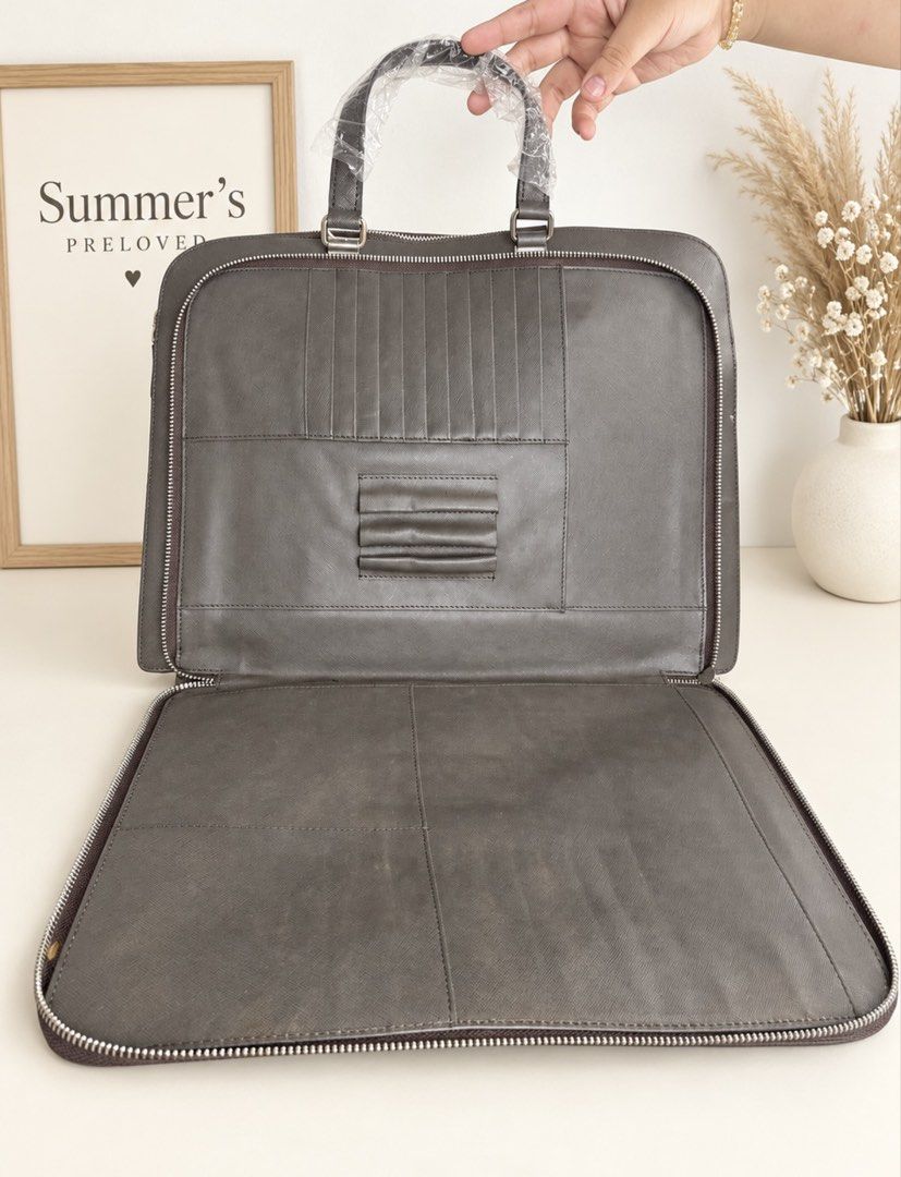 Pradudung Saffiano Leather Briefcase (Category: Laptop Bags & Sleeves) located in Taguig. Submitted by summers.preloved (Listing ID 1462232179, Image 3). Description: 👜 Bag Features 💜 Signature Saffiano crosshatch leather 🩶 Dark charcoal/graphite colour 💼 Structured briefcase silhouette 🤎 Double top handles with protective wrapping 🔗 Silver-tone hardware 🔺 Prada enamel triangle logo 🏷️ Prada-branded hanging leather tag and metal charm 🔐 